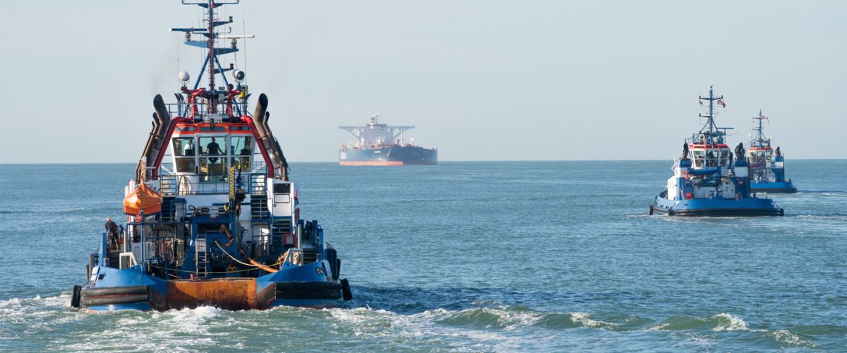 Drei Schlepper fahren auf offener See, im Hintergrund ein Frachtschiff 